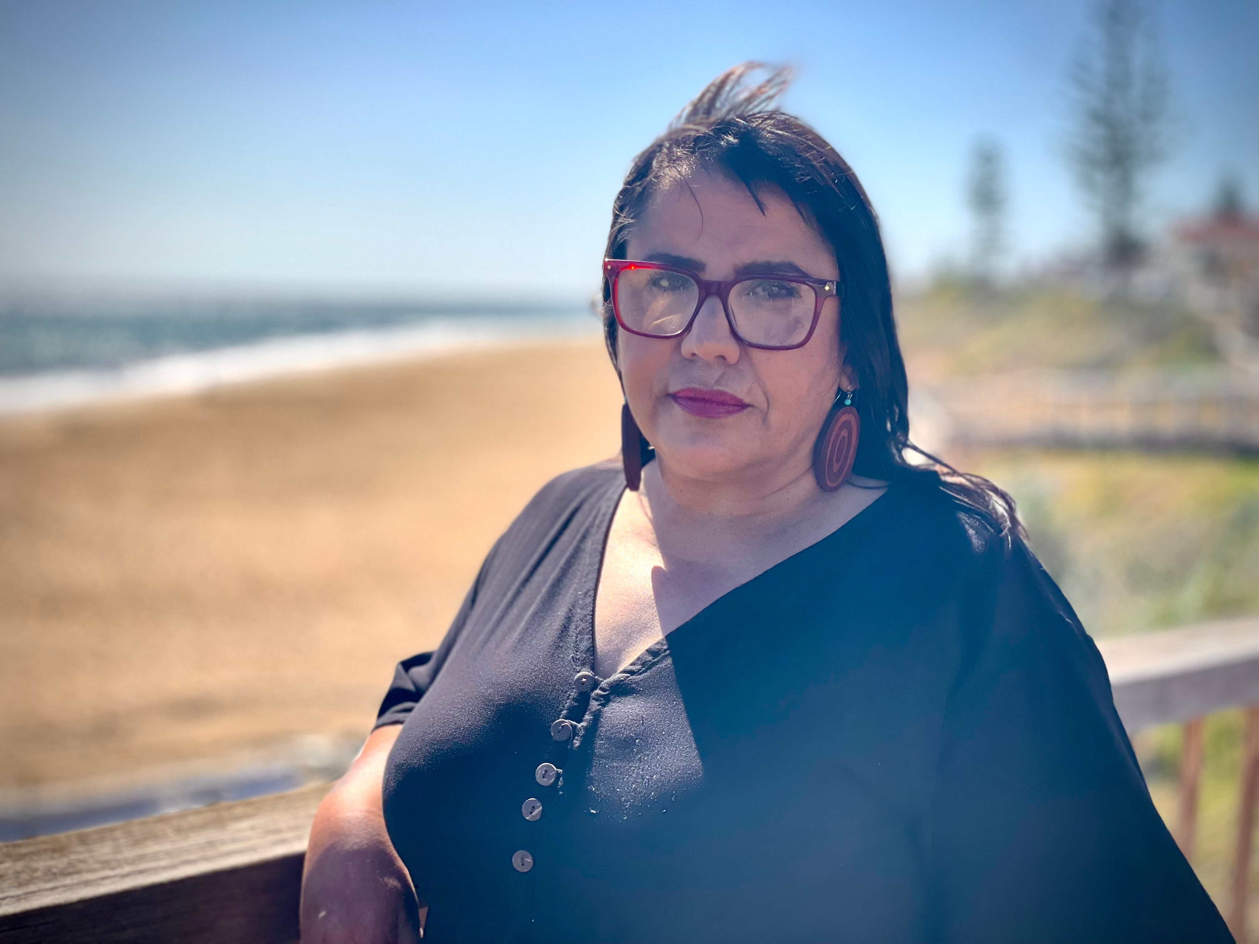 Una mujer con gafas rojas, pendientes circulares de color naranja y marrón y un top negro se apoya en un poste de madera en la playa con el codo. 