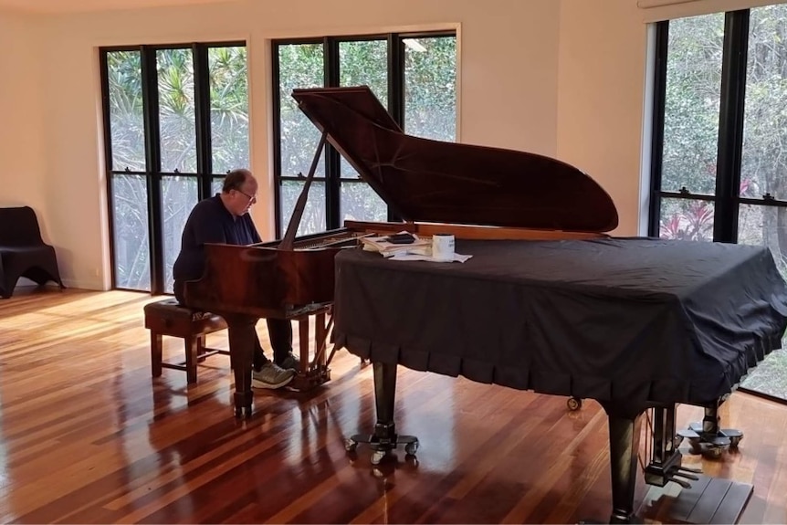 A man sitting at a grand piano inside a concert hall with wooden floorboard.