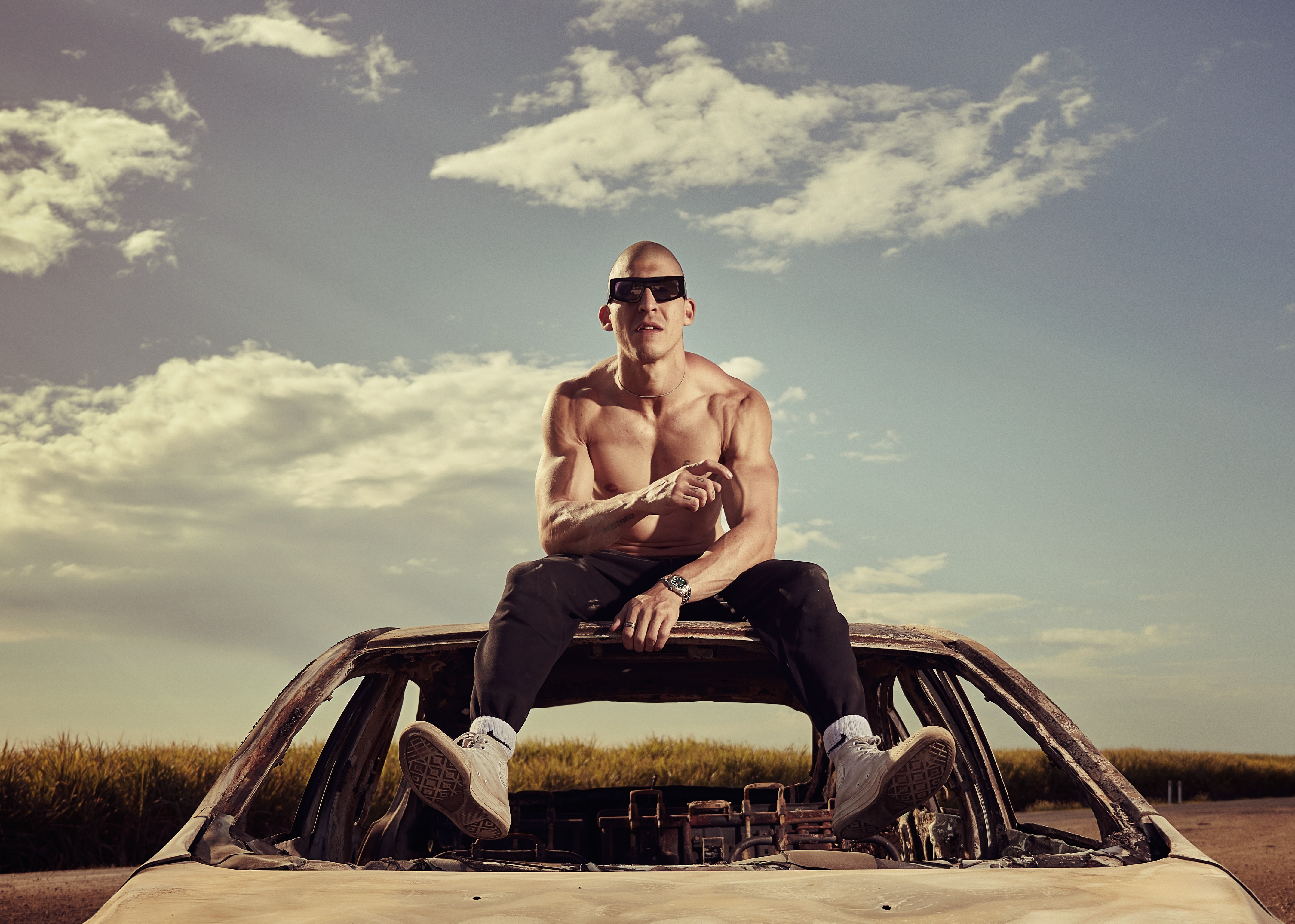 Chillinit sits on top of a car roof. He is shirtless with black sunglasses on and a shaved head.