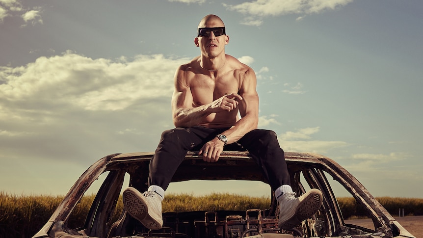 Chillinit sits on top of a car roof. He is shirtless with black sunglasses on and a shaved head.