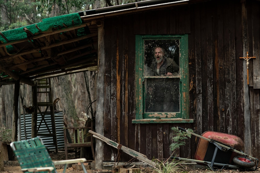 A film still of Hugo Weaving. He is inside a ramshackle house and can be seen through the window, yelling.