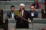 An older man with white hair wearing a suit, a purple scarf and waving a bamboo stick reads from a yellow folder in parliament.