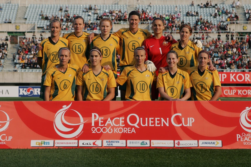 A women's soccer team wearing yellow and green poses for a photo before a game behind an advertising board
