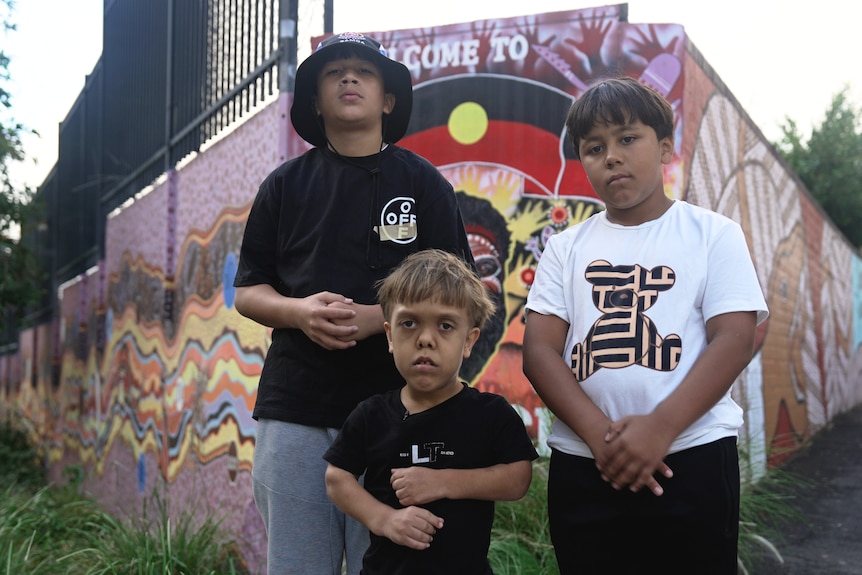 Three teenage boys, one with dwarfism, stand outside in front of a colourful mural wall