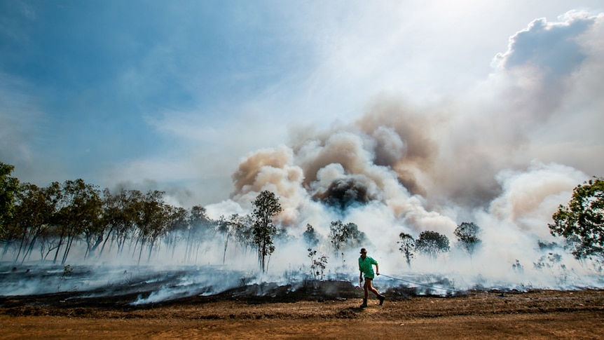 A man runs with a large hose along a dirt road in front of large clouds of smoke and smoldering ground