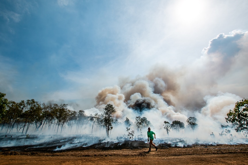 A man runs with a large hose along a dirt road in front of large clouds of smoke and smoldering ground