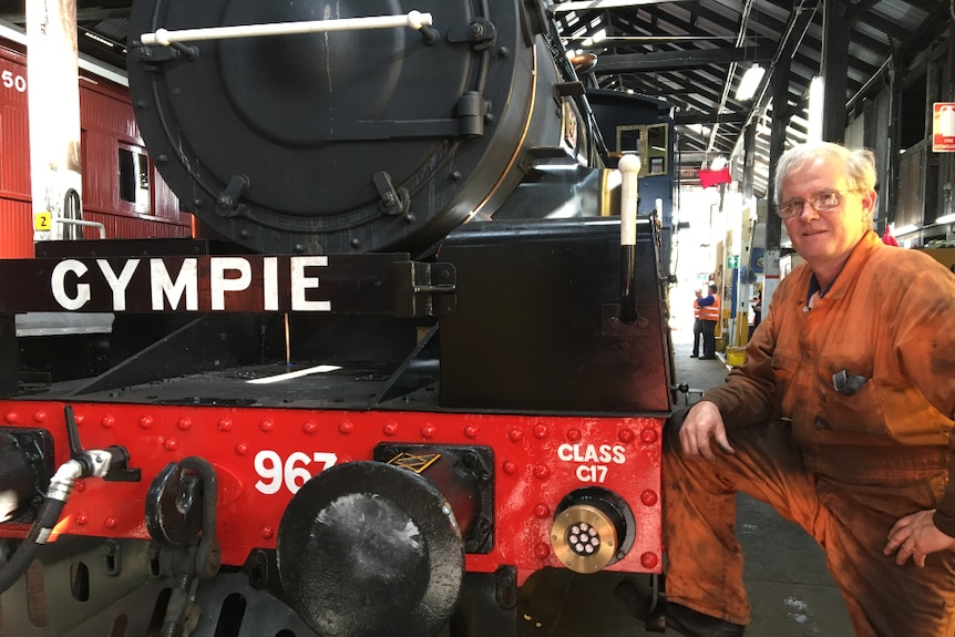 A man in dirty orange overalls stands beside a steam train in a shed.
