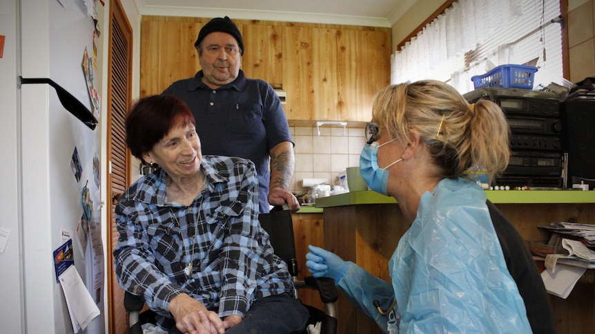 A paramedic in PPE leans down and talks to a woman in a wheelchair and her partner