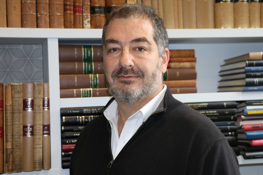 An academic-looking, middle-aged man stands in front of a bookshelf laden with leatherbound editions.