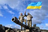 Two men dressed in army fatigues stand on a cannon while holding a ukrainian flag.