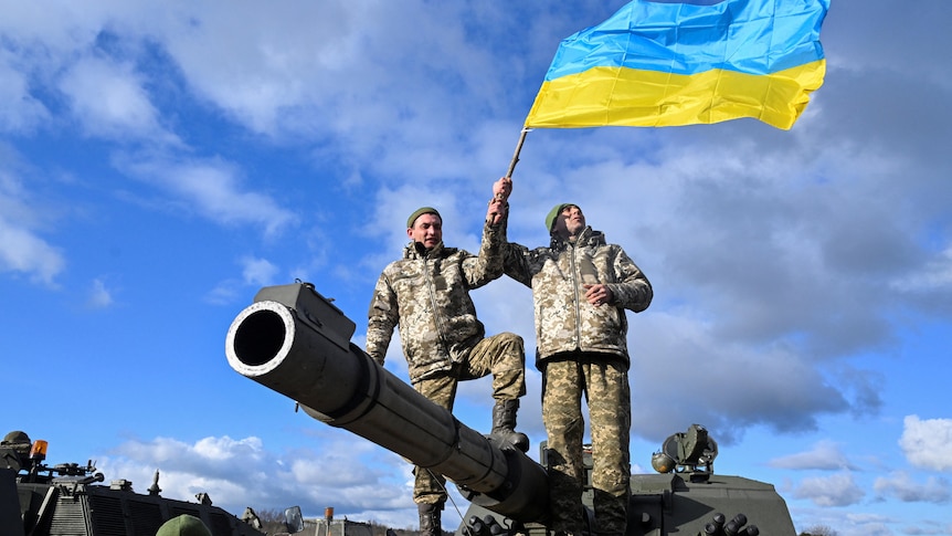 Two men dressed in army fatigues stand on a cannon while holding a ukrainian flag.