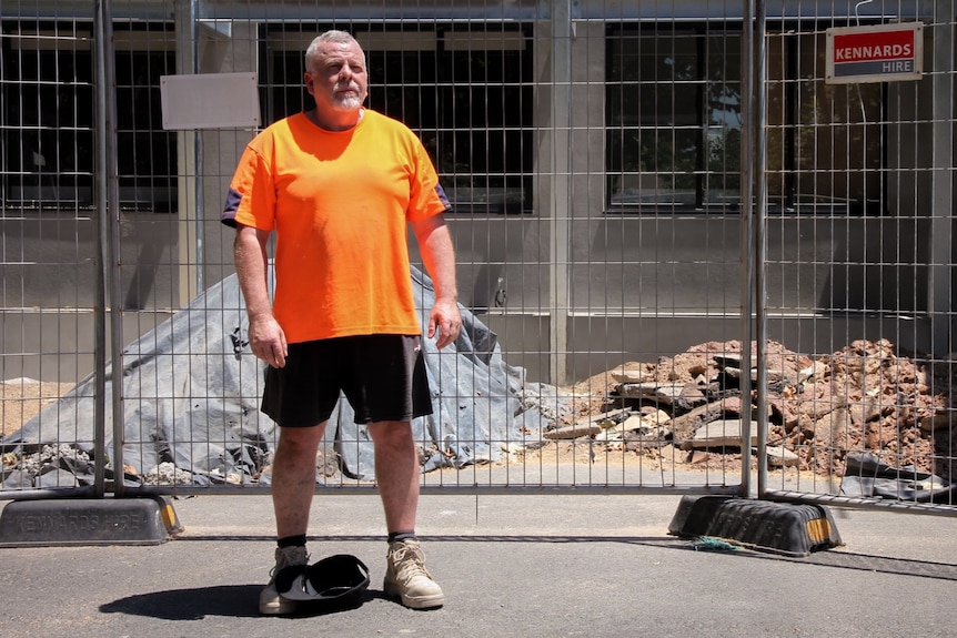 A middle-aged man in an orange hi-vis shirt standing in front of a construction site.