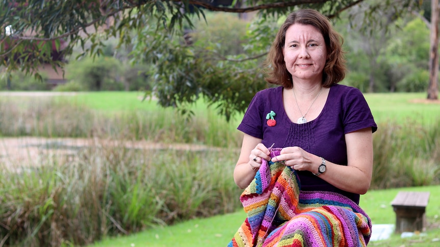 Pippa Burns works on some crochet on a park seat with some of her crocheted items on the seat next to her.