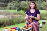 Pippa Burns works on some crochet on a park seat with some of her crocheted items on the seat next to her.