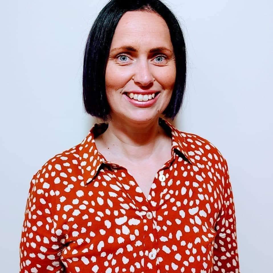 A woman with a brown bob wears a red and white spotty shirt and smiles at the camera