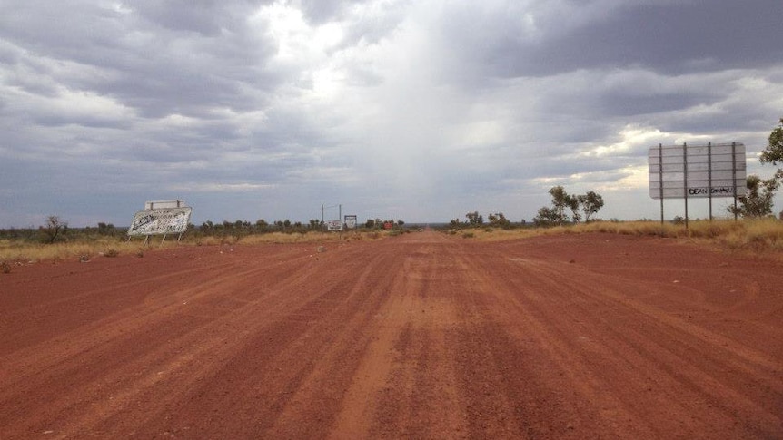 The red gravel Tanami track stretches into the distance under a grey clouded sky.