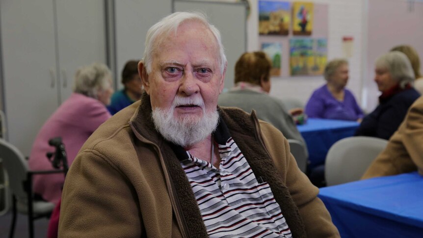 A man with white hair and dark circles under his eyes looks directly at the camera.