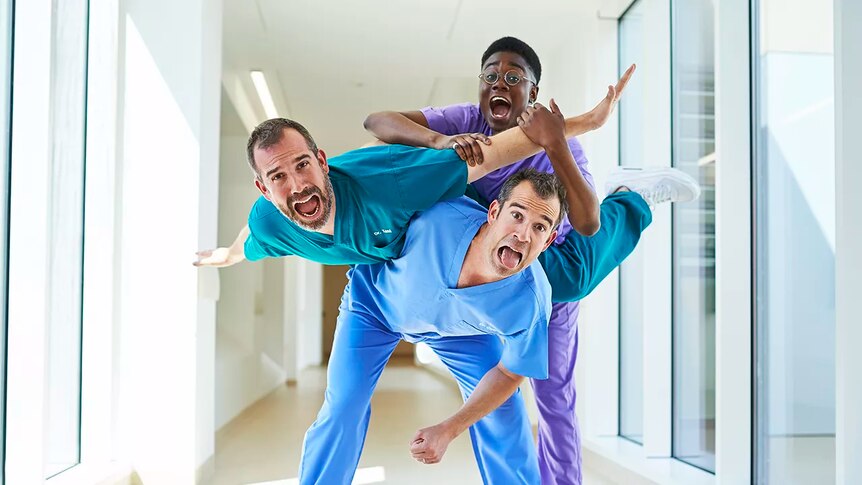 Three people, two of them twins, in hospital scrubs in an exhuberant human pyramid