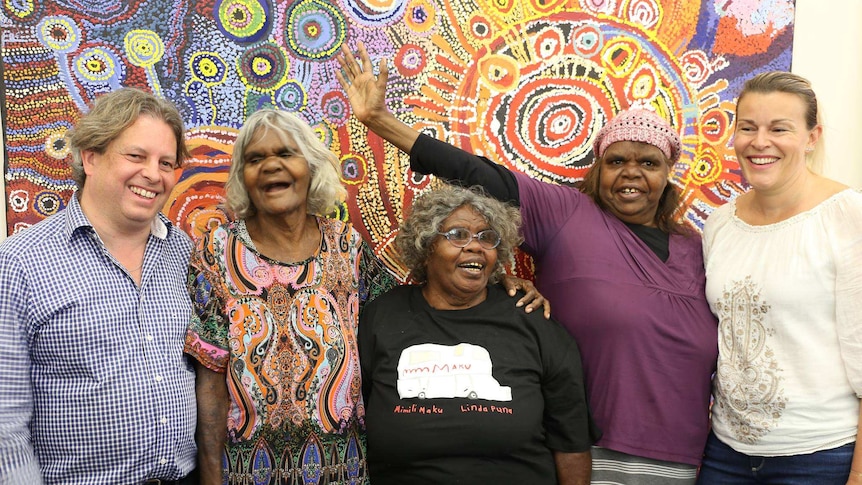 Three Aboriginal female elders stand with two white art collectors smiling in front of large traditional Aboriginal painting