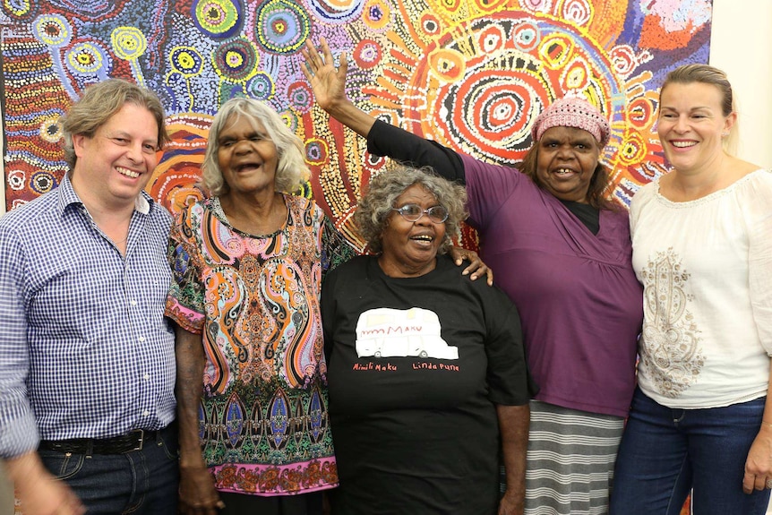 Three Aboriginal female elders stand with two white art collectors smiling in front of large traditional Aboriginal painting