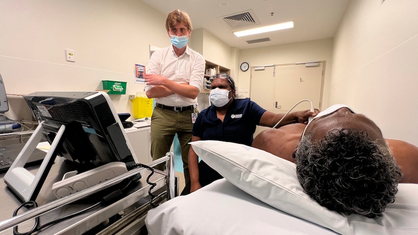 A man in a white shirt with arms folded watches a computer as a nurse holds an ultrasound prob on a man's chest.