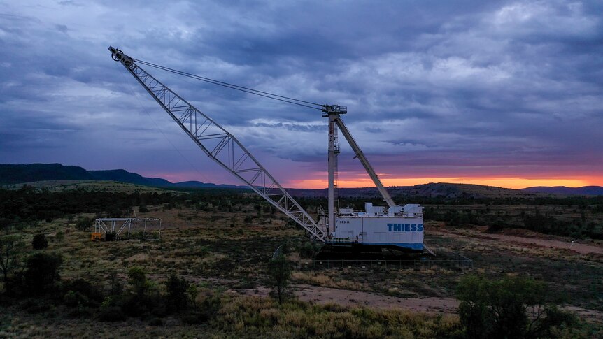 Large white dragline excavator machine on patchy landscape with sunset and dark blue clouds in background