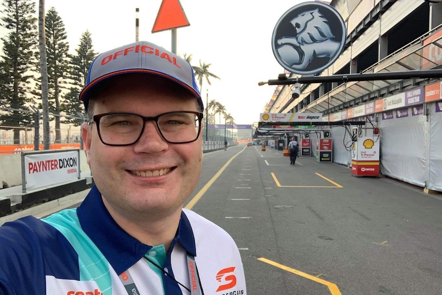 A man wearing a t-shirt and hat with racing logos stands near a street circuit.