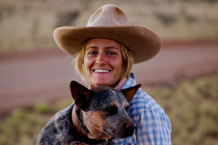 A woman wearing a large hat sits with a blue heeler dog in her arms