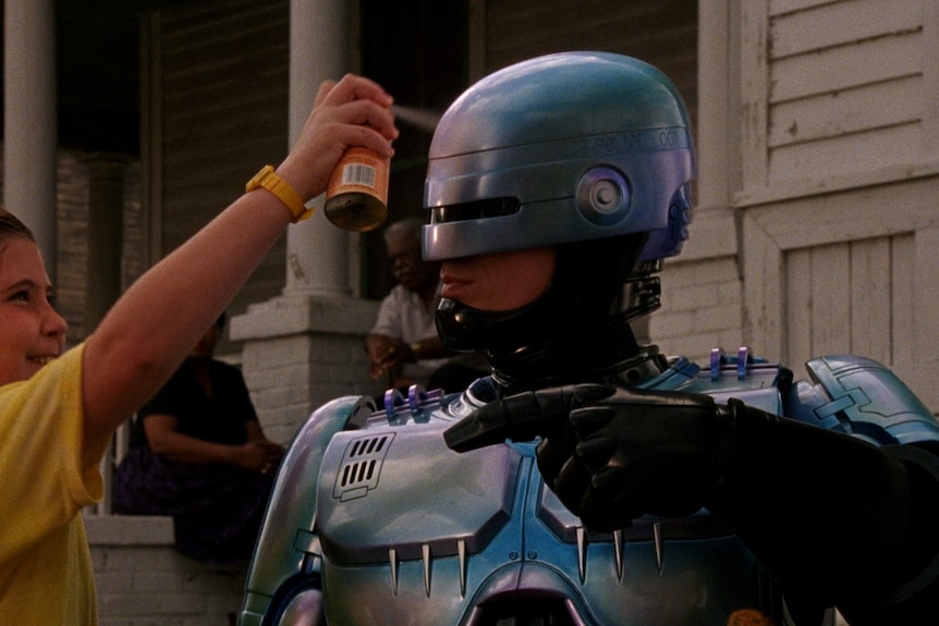 A film still from RoboCop 2, of a man in a robotic costume, his mouth visible, as a little girl sprays a can on his helmet