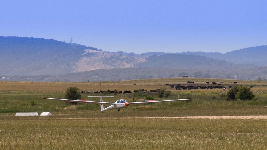 A white and red glider landing in front of paddocks and hills