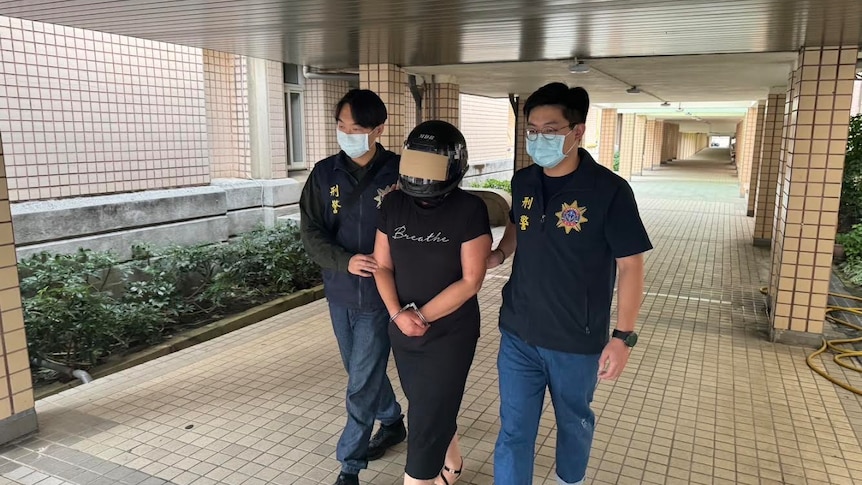 A woman wearing a helmet is escorted by two men wearing face masks outside a police station.