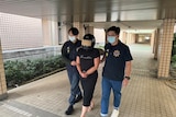 A woman wearing a helmet is escorted by two men wearing face masks outside a police station.