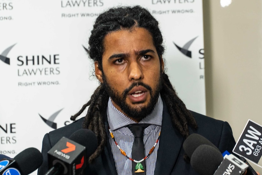 A man stands in front of a row of microphones in a law office as he answers reporters' questions.