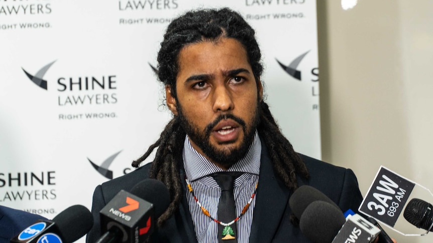 A man stands in front of a row of microphones in a law office as he answers reporters' questions.