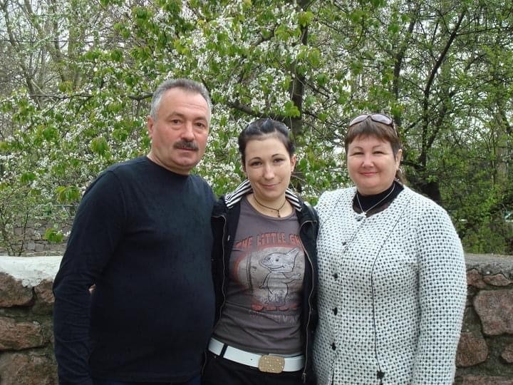 A man and woman stand with a younger woman in between them, in front of trees