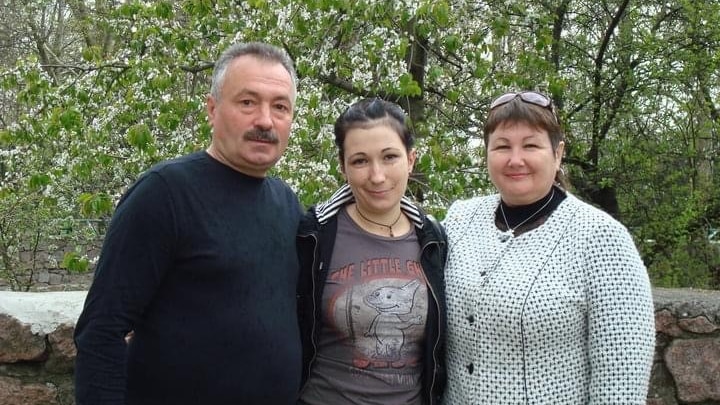 A man and woman stand with a younger woman in between them, in front of trees