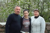 A man and woman stand with a younger woman in between them, in front of trees