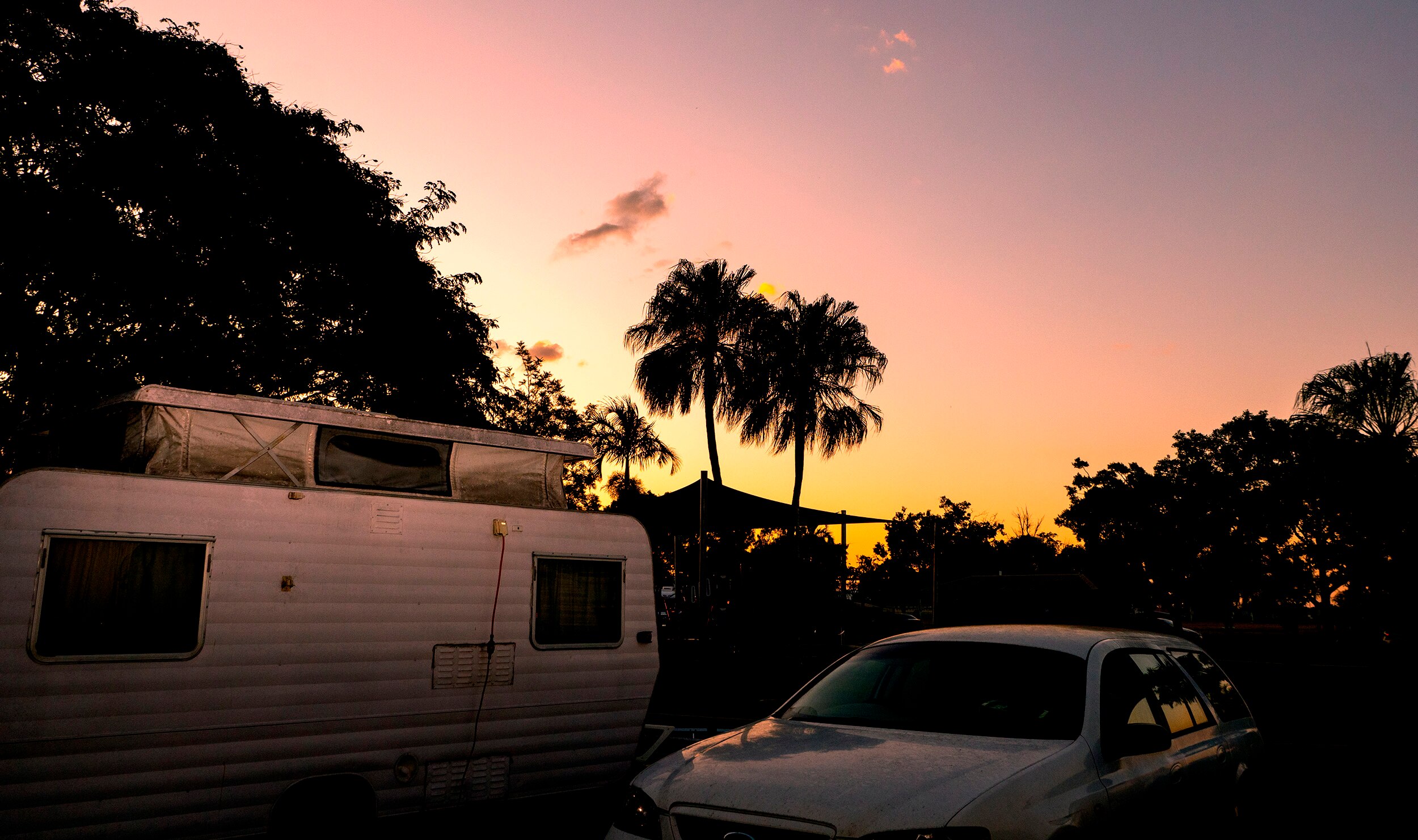 A car and caravan with tropical sunset, palm trees in the background.