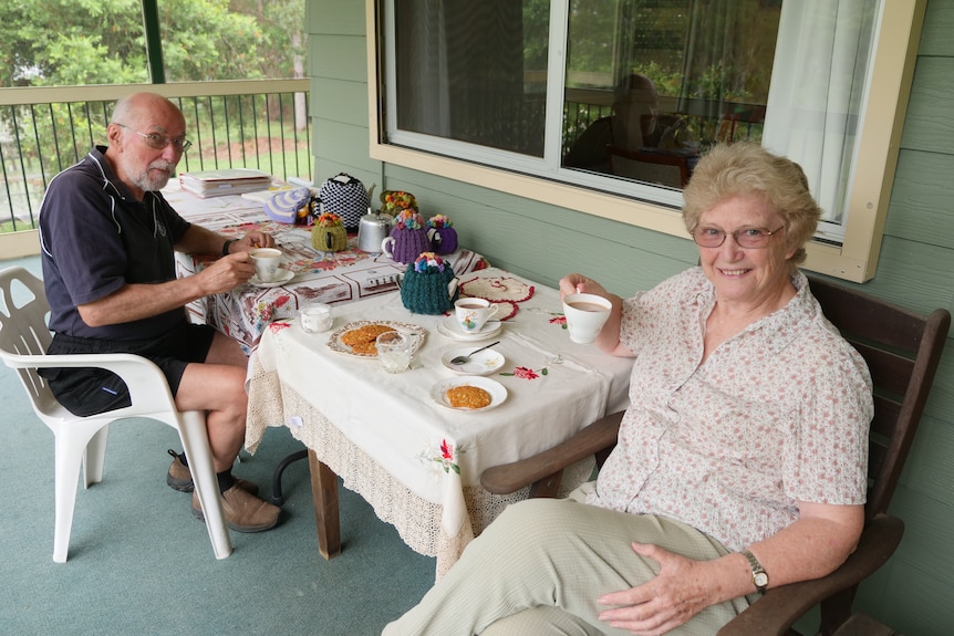 An older man and woman sit on a deck drinking a cup of tea, with a teapot on the table.