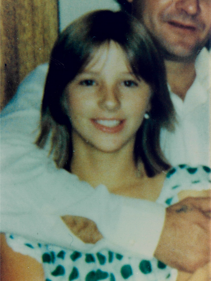 A young girl smiles at a camera, with a man's arm around her.