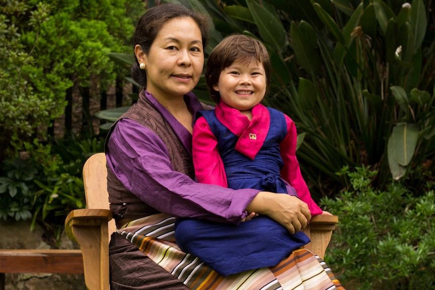 Kyinzom Dhongdue sitting, in traditional Tibetan clothing, with her daughter on her knee and garden behind them.