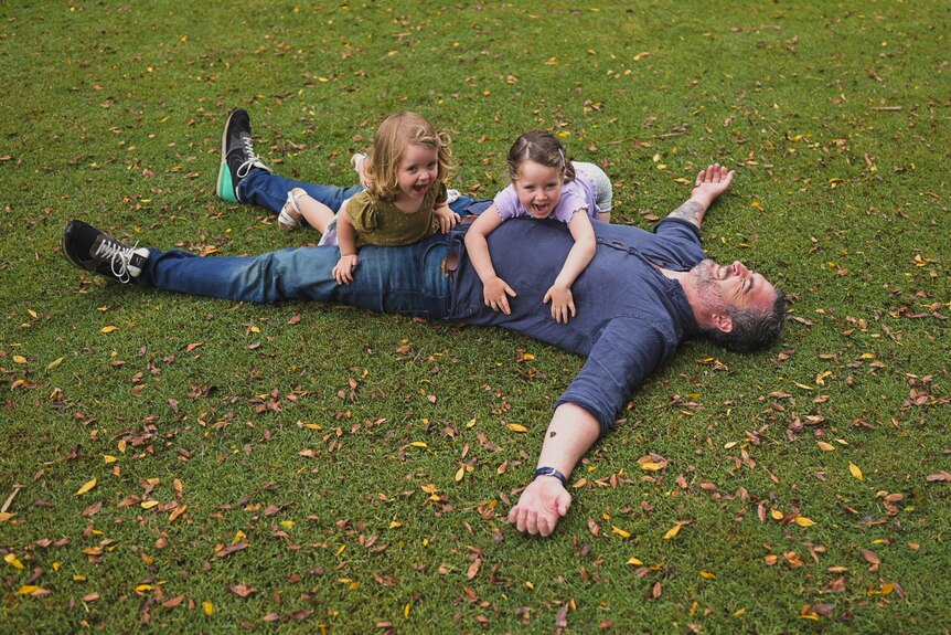 A man lies on his back with two little girls climbing on him.