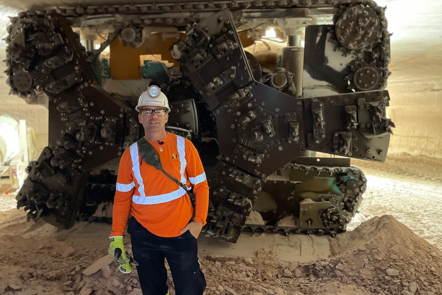 A man wearing a high vis shirt and mining helmet stands in front of a stationary piece of mining equipement