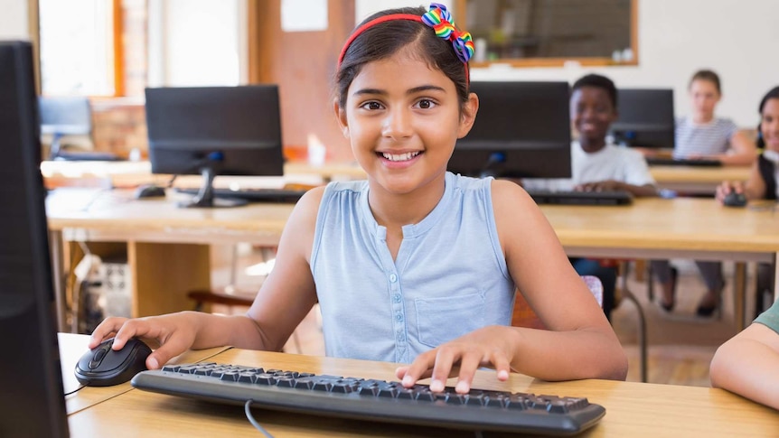 A smiling primary-school-aged girl is sitting at a computer in a classroom. Other students are sitting at desks behind her.