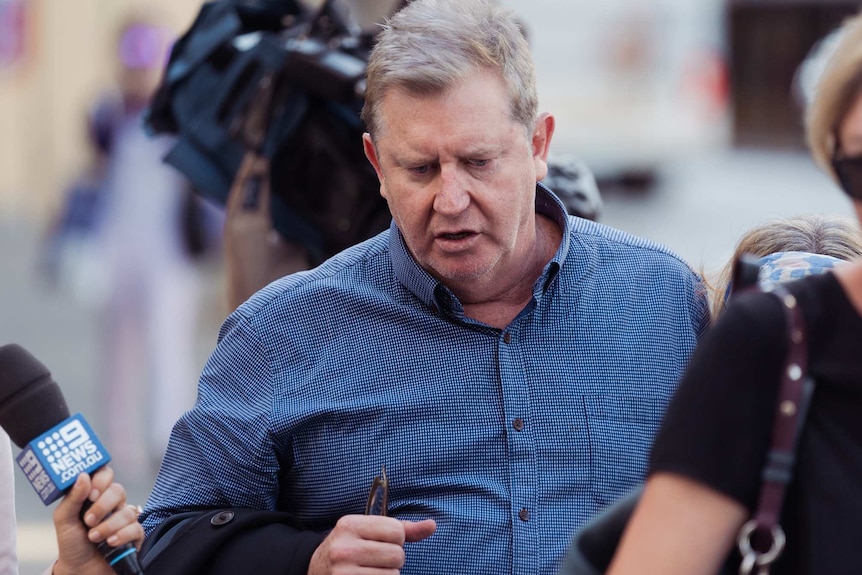A man in a blue shirt is questioned by reporters as cameras film him on the street.