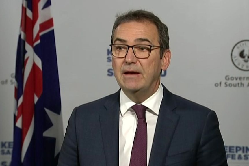 A late middle-aged man, wearing glasses and a suit, stands in front of a flag at a press conference.