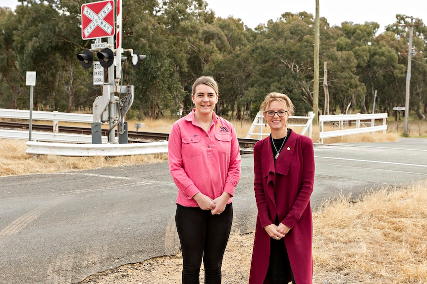 A woman in a pink shirt and a woman wearing a red coat stand on the side of a road. behind them a sign read "rail crossing, sto