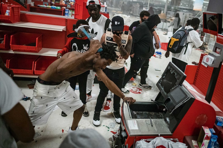 A shirtless man smashes a Target self-serve cash register with a hammer. Other people watch on.