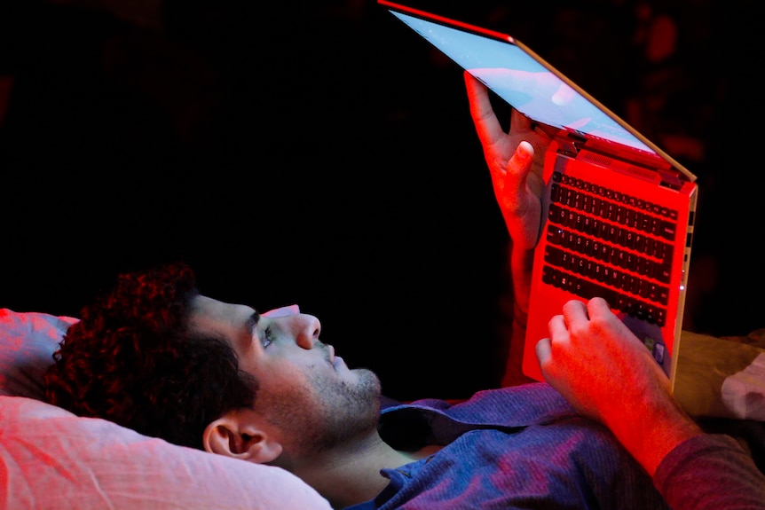 A South Asian man lies on a bed, looking at a laptop that he rests on his chest