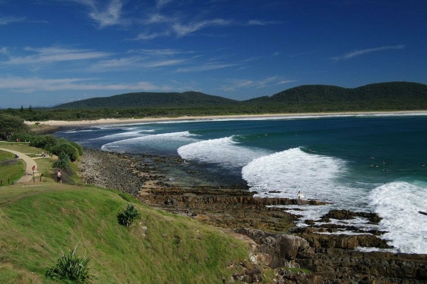 Waves roll in around a coastal headland on a sunny day.